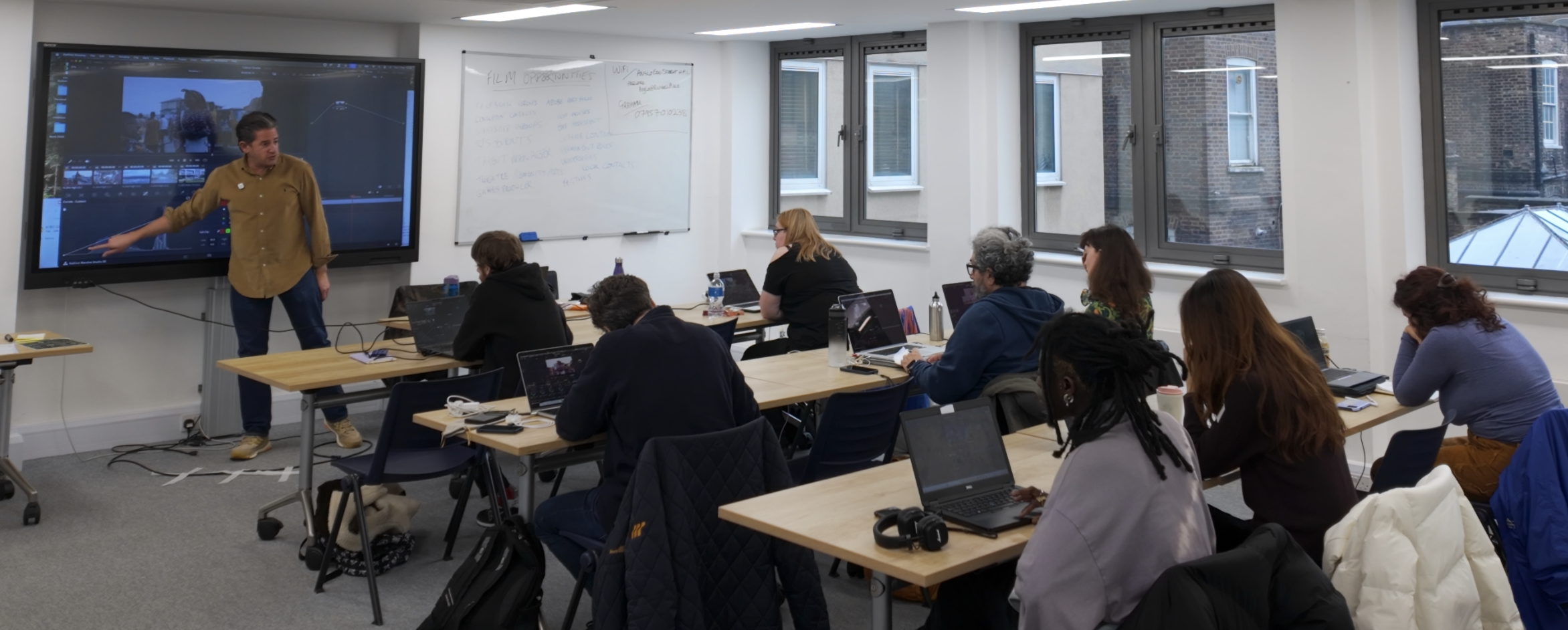 A classroom or workshop with a instructor at the front pointing to a large screen displaying video editing software, and adult students at desks with laptops, notebooks, and drinks, in a room with white walls and windows.