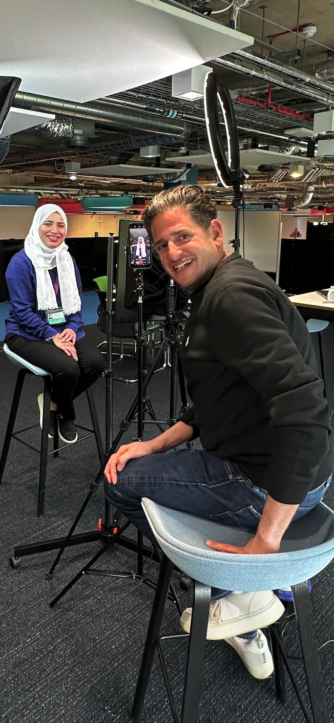 A woman wearing a white hijab and a man in casual black clothing are filming a video in a modern office space with exposed ceilings. The woman sits on a high stool and smiles at the camera, while the man crouches beside a professional camera setup, smiling at the camera. There is a ring light and a smartphone mounted on the camera setup, capturing the woman's video.