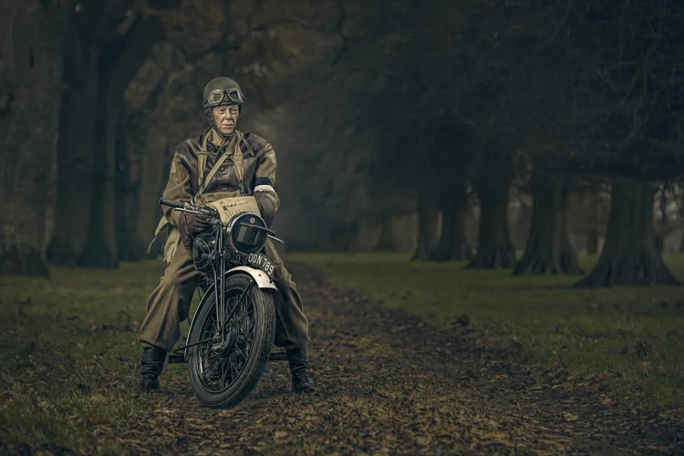 Soldier in a Second World War-style uniform on a vintage motorbike in a tree-lined avenue