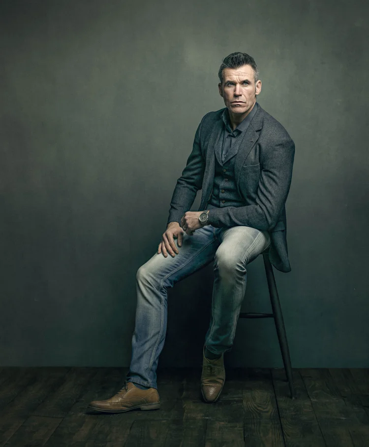 Man in a dark jacket and jeans sitting on a stool, studio portrait