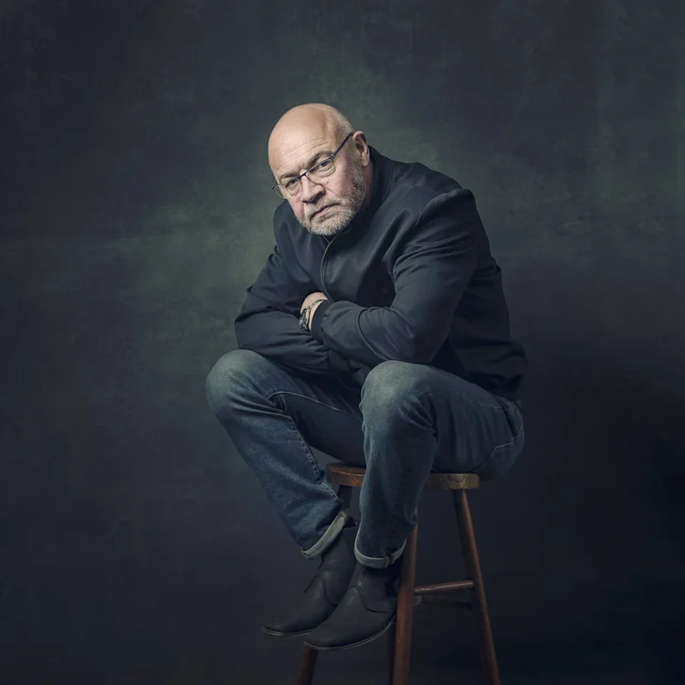 Older man in a dark jacket sitting on a stool against a mottled backdrop
