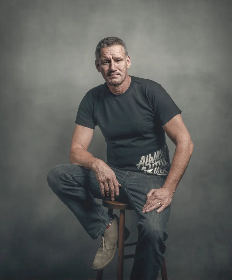 Man in a T-shirt and jeans sitting on a stool against a grey backdrop