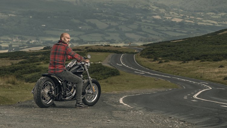 Man in a red checked shirt riding a motorbike along a winding coastal road