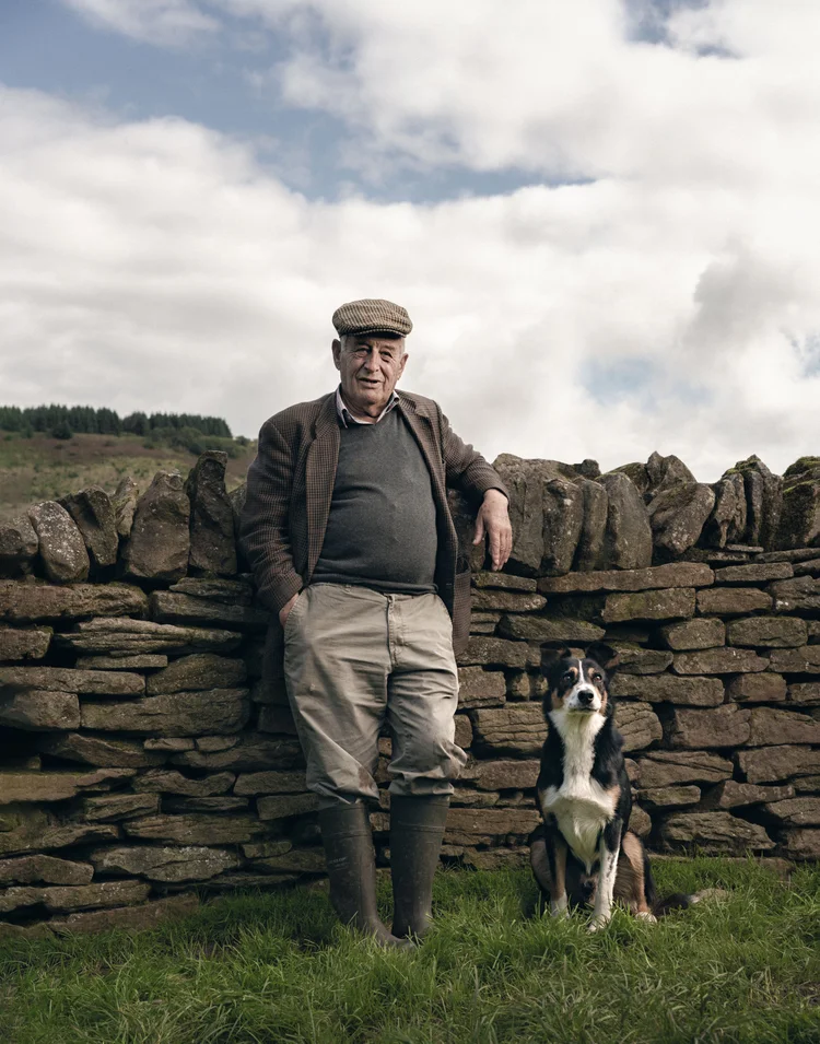 Farmer in a flat cap leaning on a dry stone wall with his sheepdog