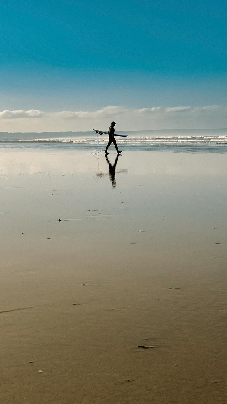 Person walking across wet sand with their reflection at Saunton