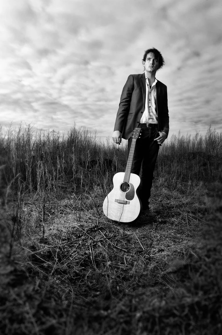 Black and white portrait of a man in a suit holding an acoustic guitar in a field