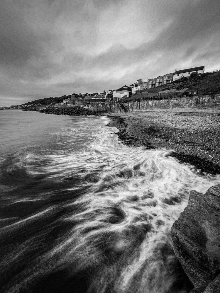 Black and white long exposure of waves washing up a shingle beach below a hillside town