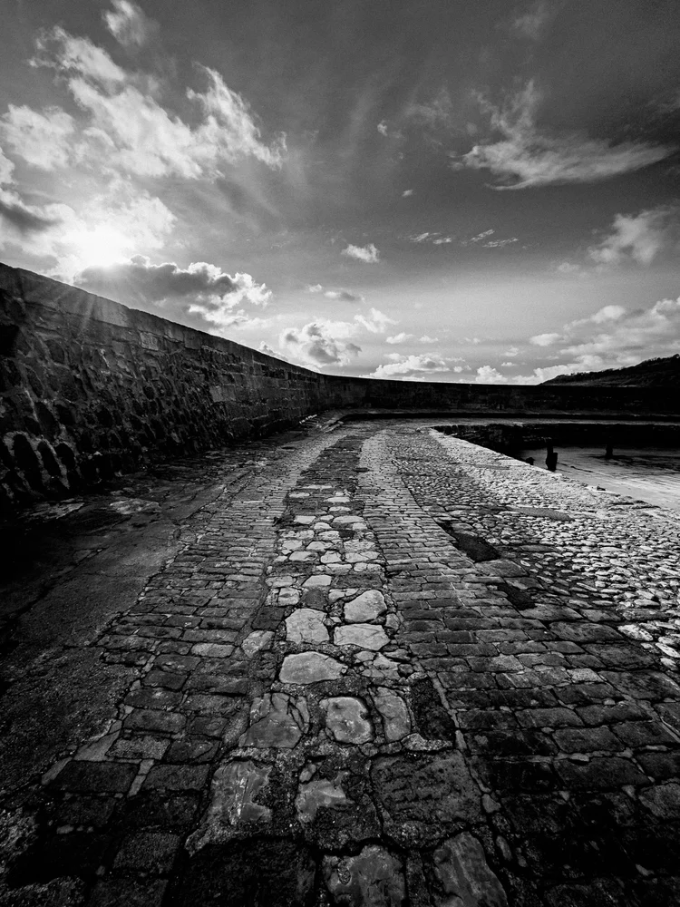 Black and white image of the stones of The Cobb leading out to sea under a dramatic sky