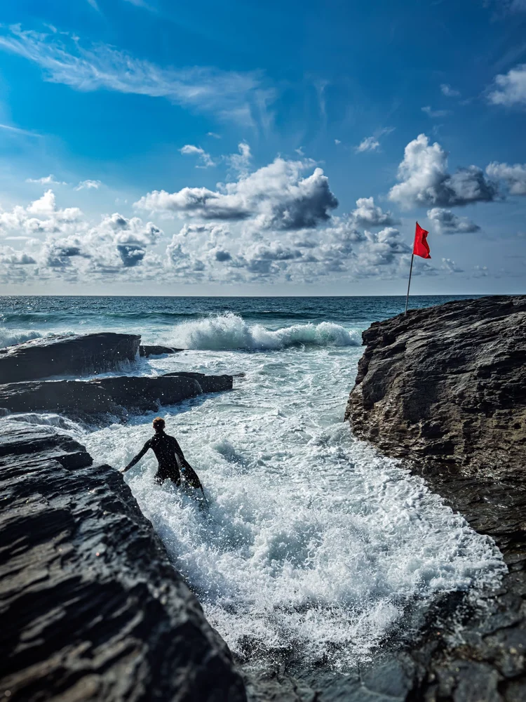 Surfer heading out between rocks and breaking waves under a blue sky