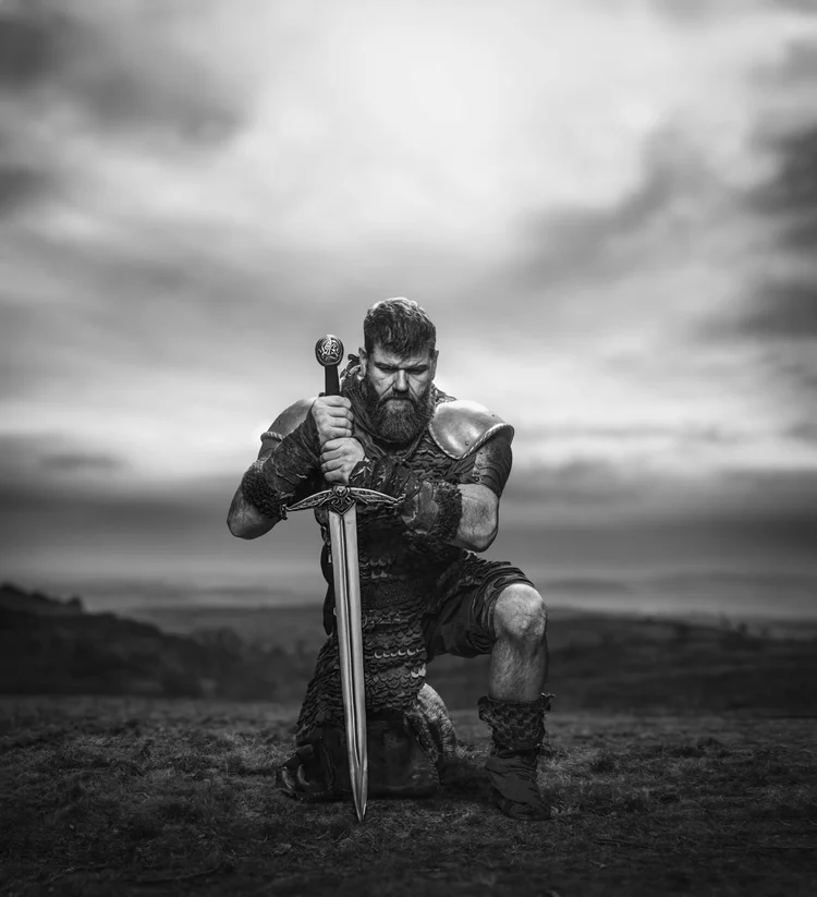 Black and white portrait of a warrior kneeling with a sword on a hillside