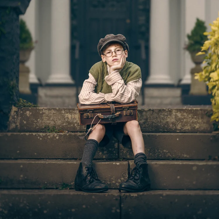 Boy in a flat cap sitting on the steps of a grand house with a small suitcase