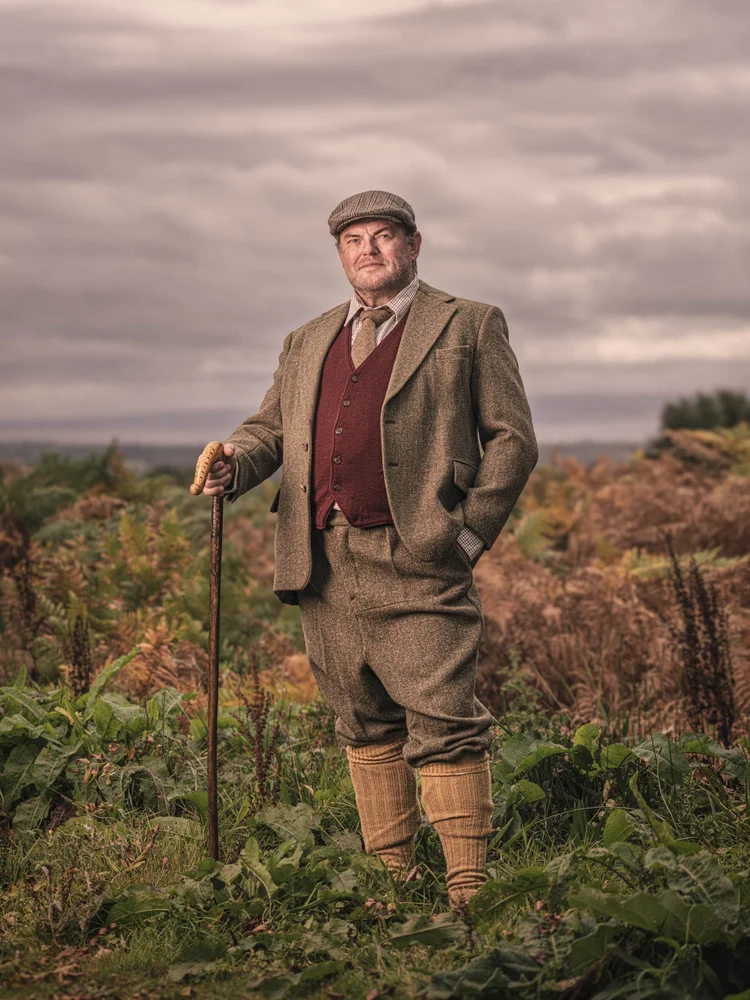 Man in a tweed suit and flat cap with a walking stick in open countryside