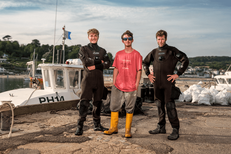 Three divers in drysuits on the quayside in front of a fishing boat