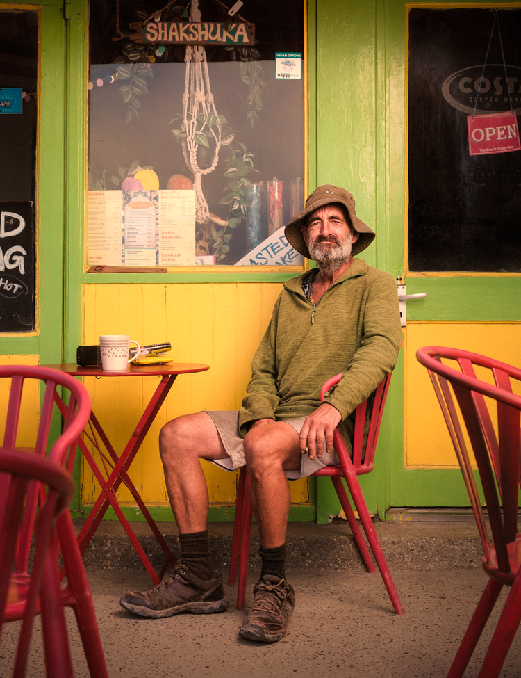 Man in a bush hat and green jacket sitting on a red chair outside a brightly painted café