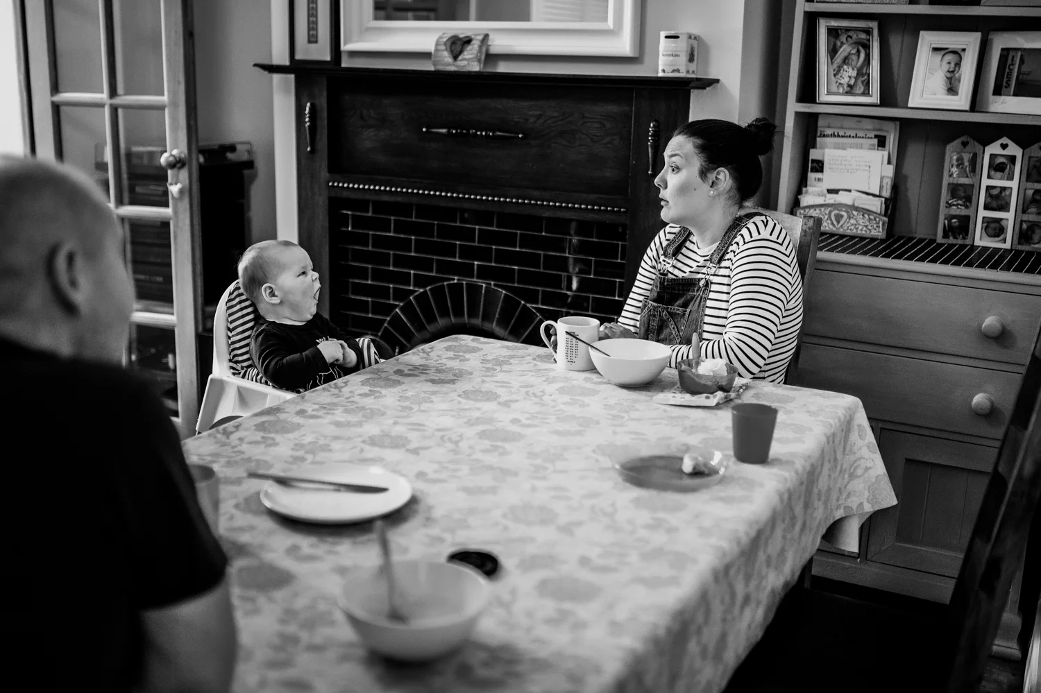 Family sit down at the table having breakfast and the baby yawns at the mother who makes a funny face back during their Day in The Life session in Bristol.