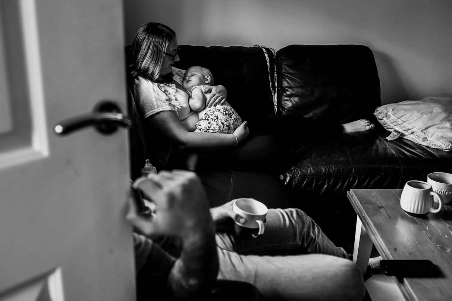 A shot through the door as her mother sits on her sofa holding her baby on her chest whilst her baby sleeps during this relaxed family photo session in Bath.