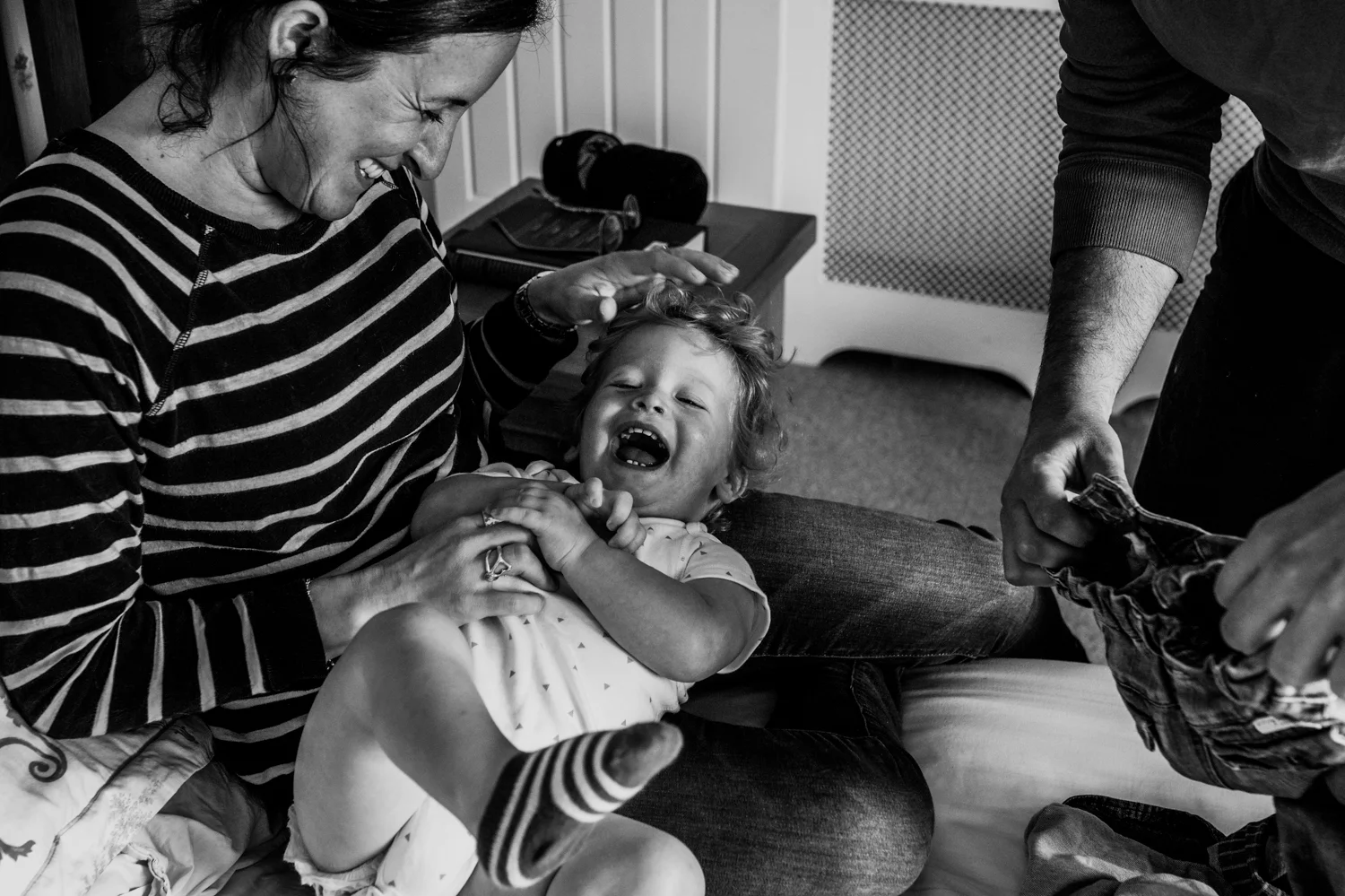 Mother tickles her toddler son on her lap whilst trying to get him dressed during in-home session in Chew Magna, Bristol.