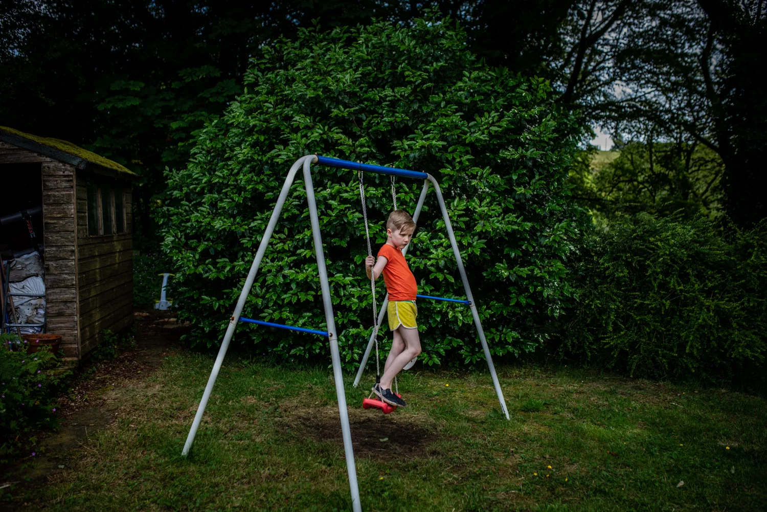 A portrait of a young boy standing on a swing in his garden, staring into the distance during this relaxed family portrait photo shoot in Manchester, UK.