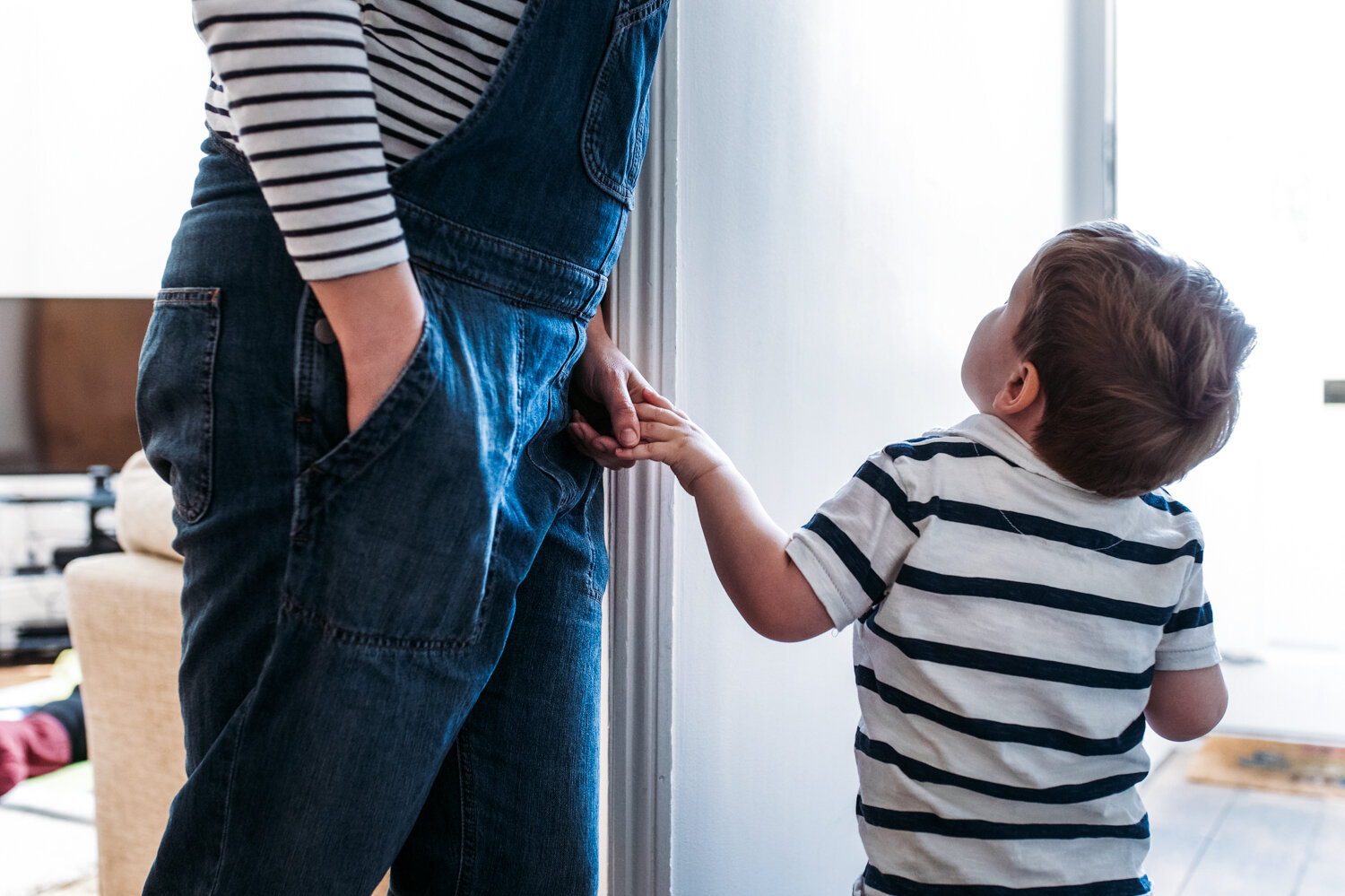  Toddler height shot of mother leaning aginst the wall in her hallway holding hands with her three year old son whilst he looks up to her during photoshoot in Bath. 