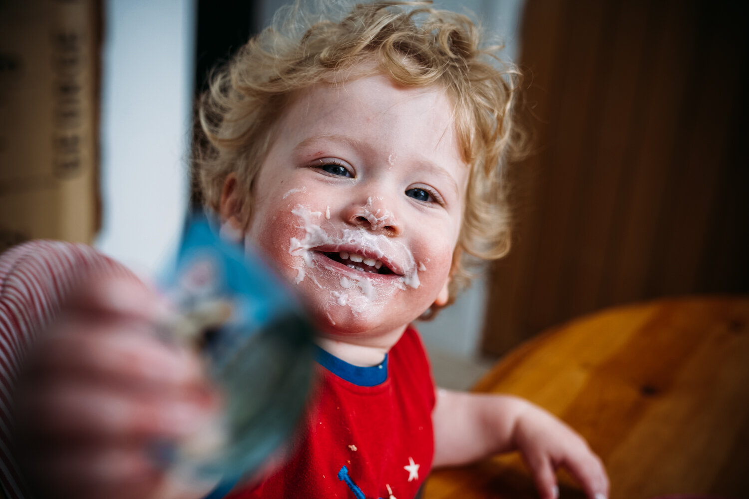  Cute toddler with curly blonde hair smiles at the camera with yoghurt round his mouth eating his breakfast during in-home session in Chew Magna, Bristol. 