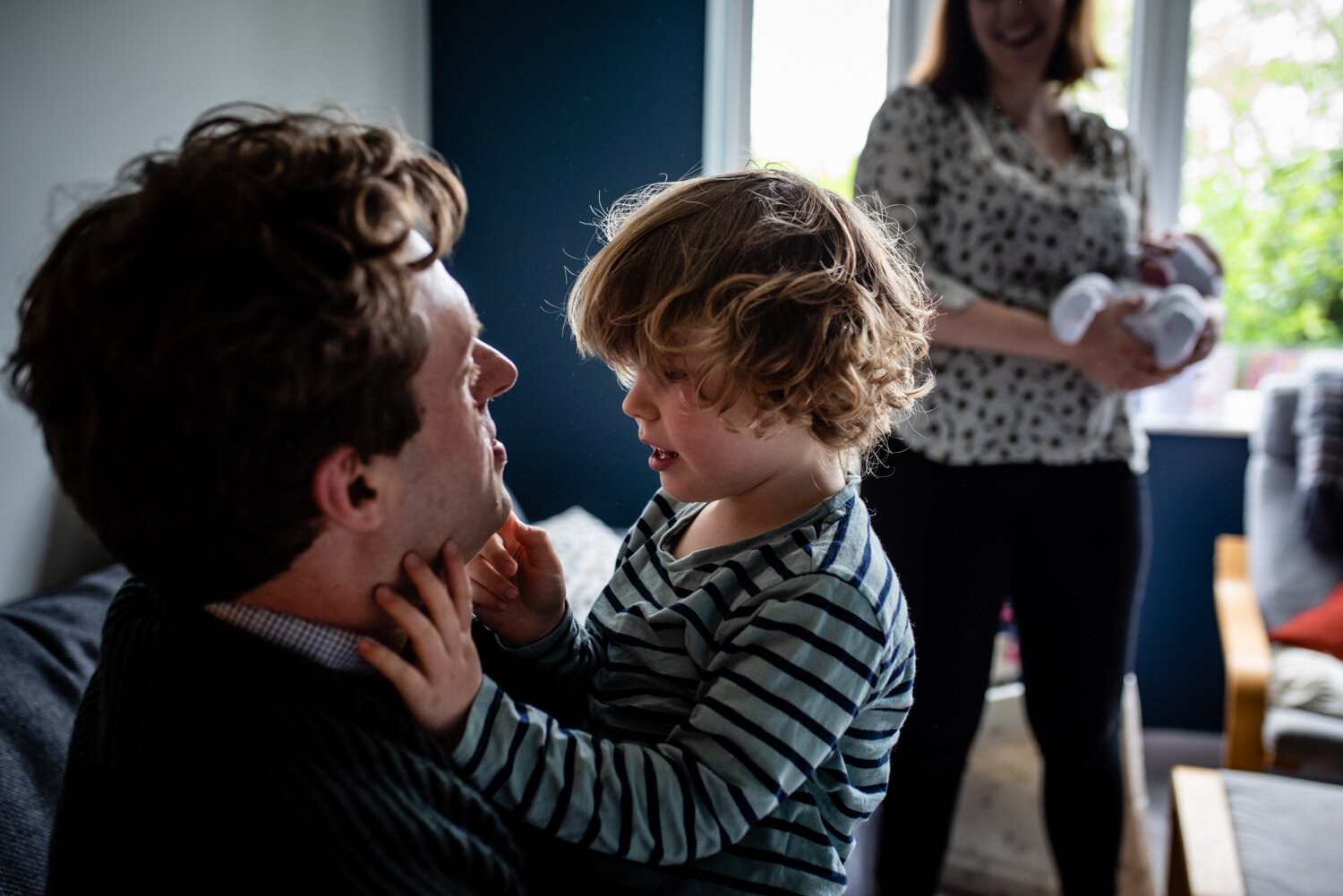  A toddler boy sits on his fathers lap holding his face and gazing into his eye during this at home documentary family photography session in Bristol. 