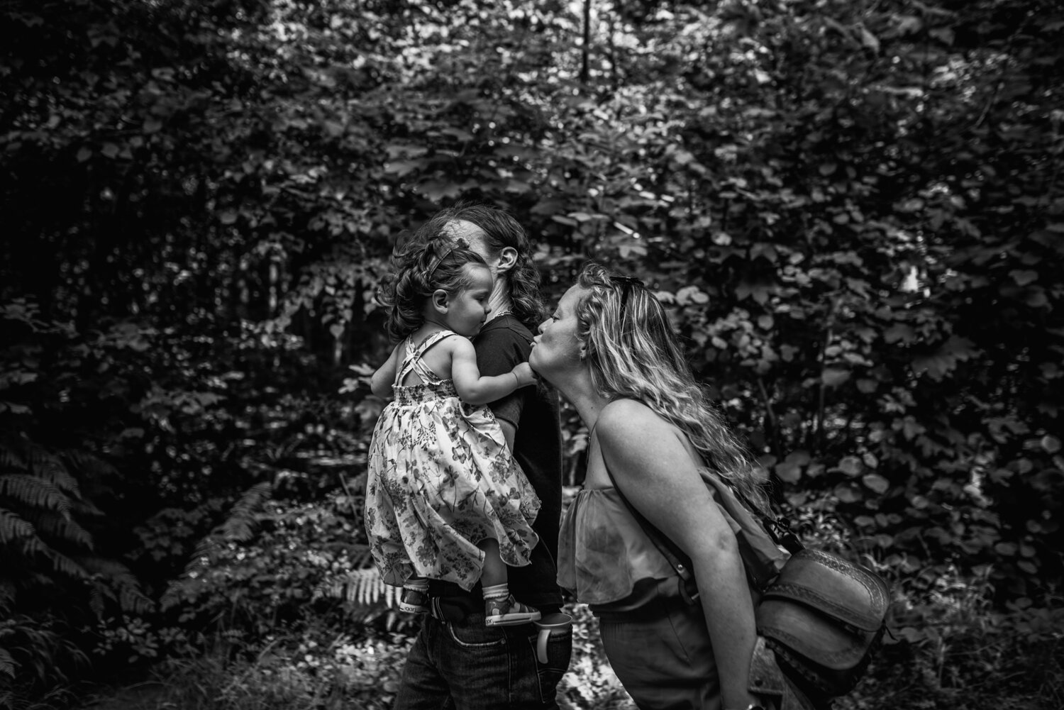  A mother leans in to kiss her daughter who is being held over her father's shoulder during this outdoor documentary family photo shoot in Leigh Woods in Bristol. 