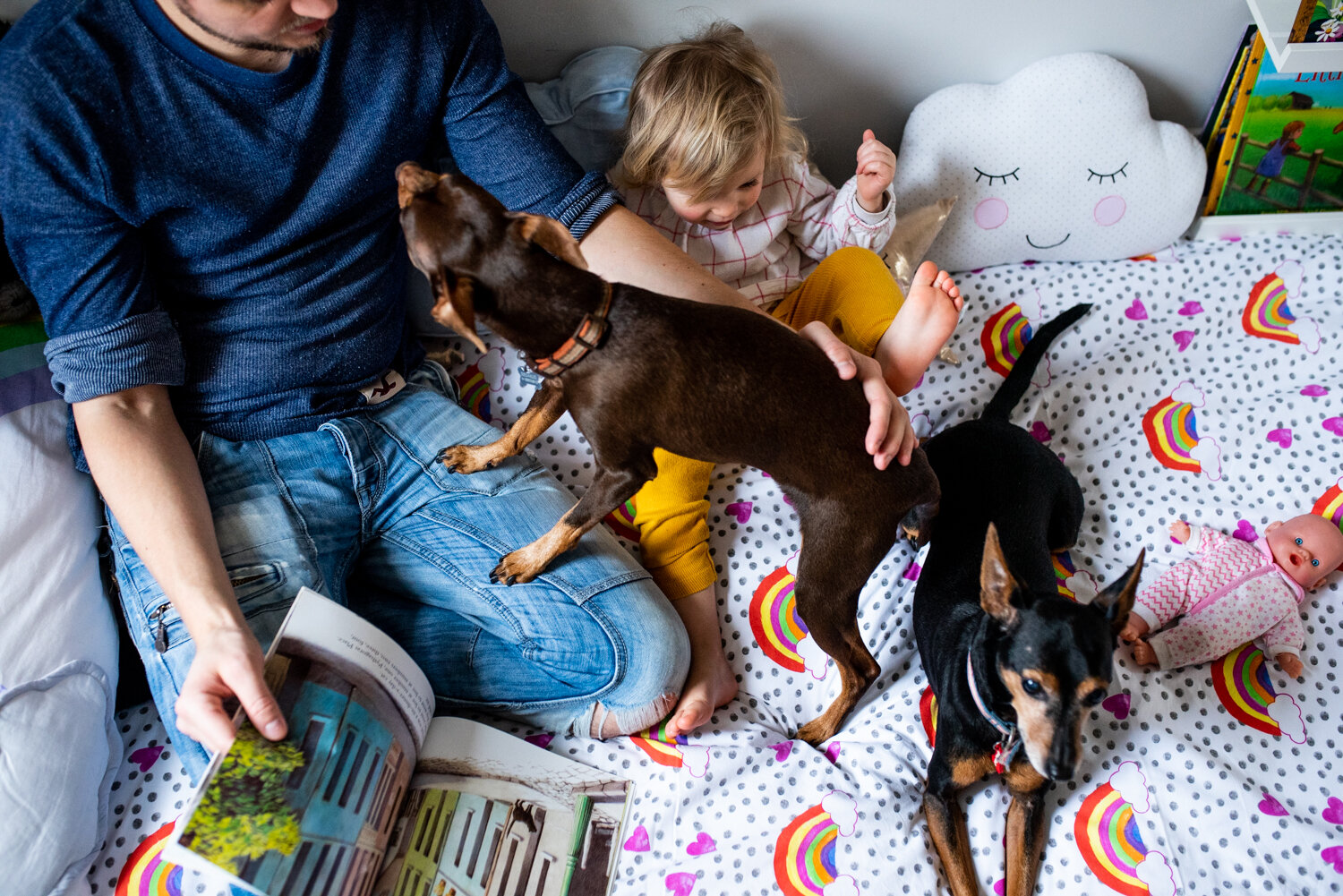  A baby girl sits on her bed next to her father who is reading her a book and they are joined by their pet dogs during this family photography session in Bath. 