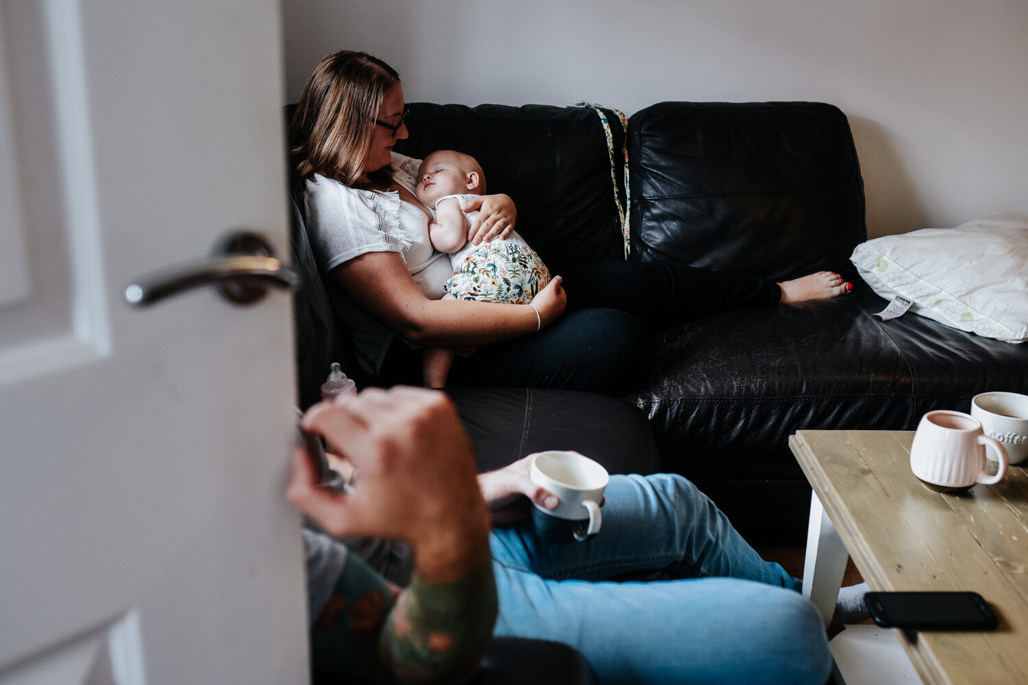  A shot through the door as her mother sits on her sofa holding her baby on her chest whilst her baby sleeps during this relaxed family photo session in Bath. 