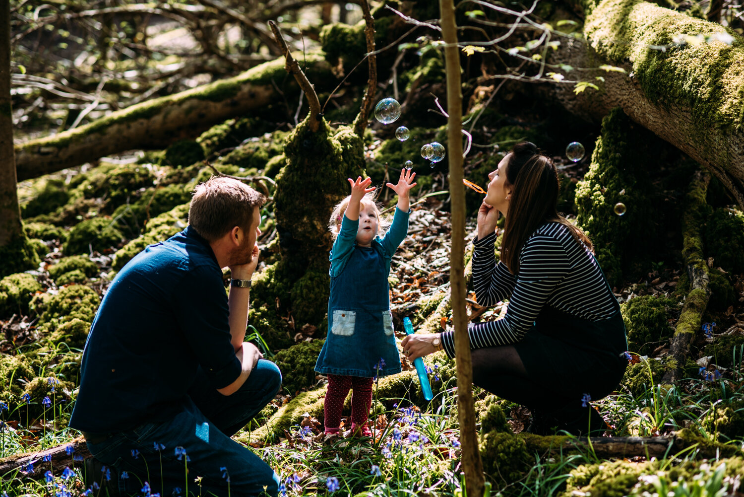  A little girl reaches up to touch bubbles her mother is blowing as the family sit amongs bluebells in a wood near Bath Somerset for this relaxed, unposed family photoshoot in Bath, Somerset. 