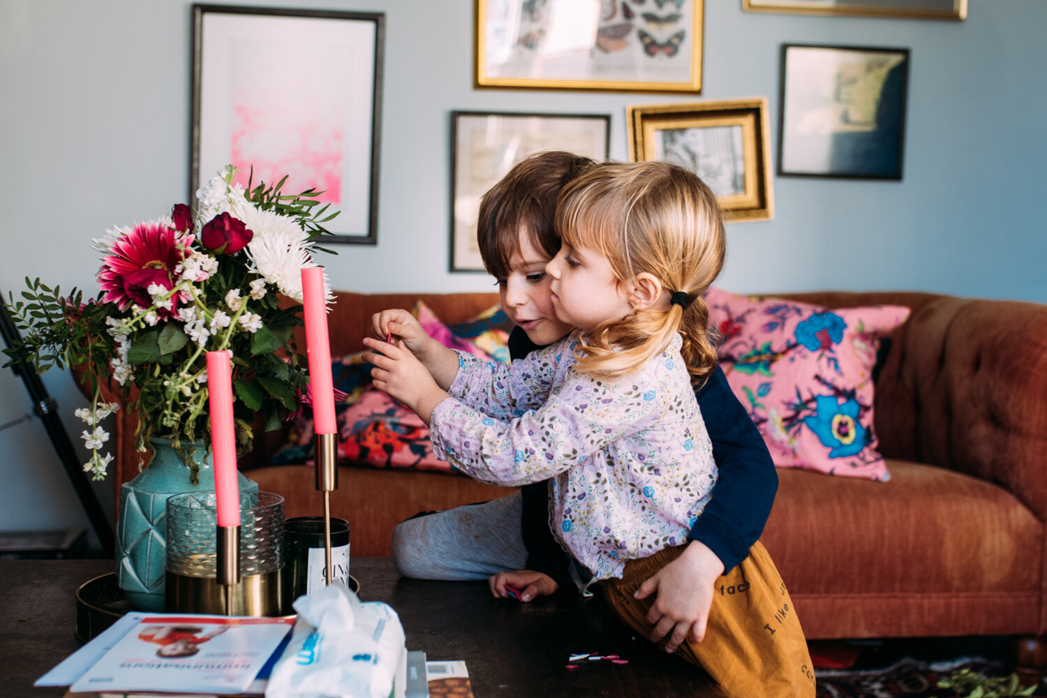  A young brother and sister stand in their living room looking at a bouquet of flowers with their arms round eachother. 
