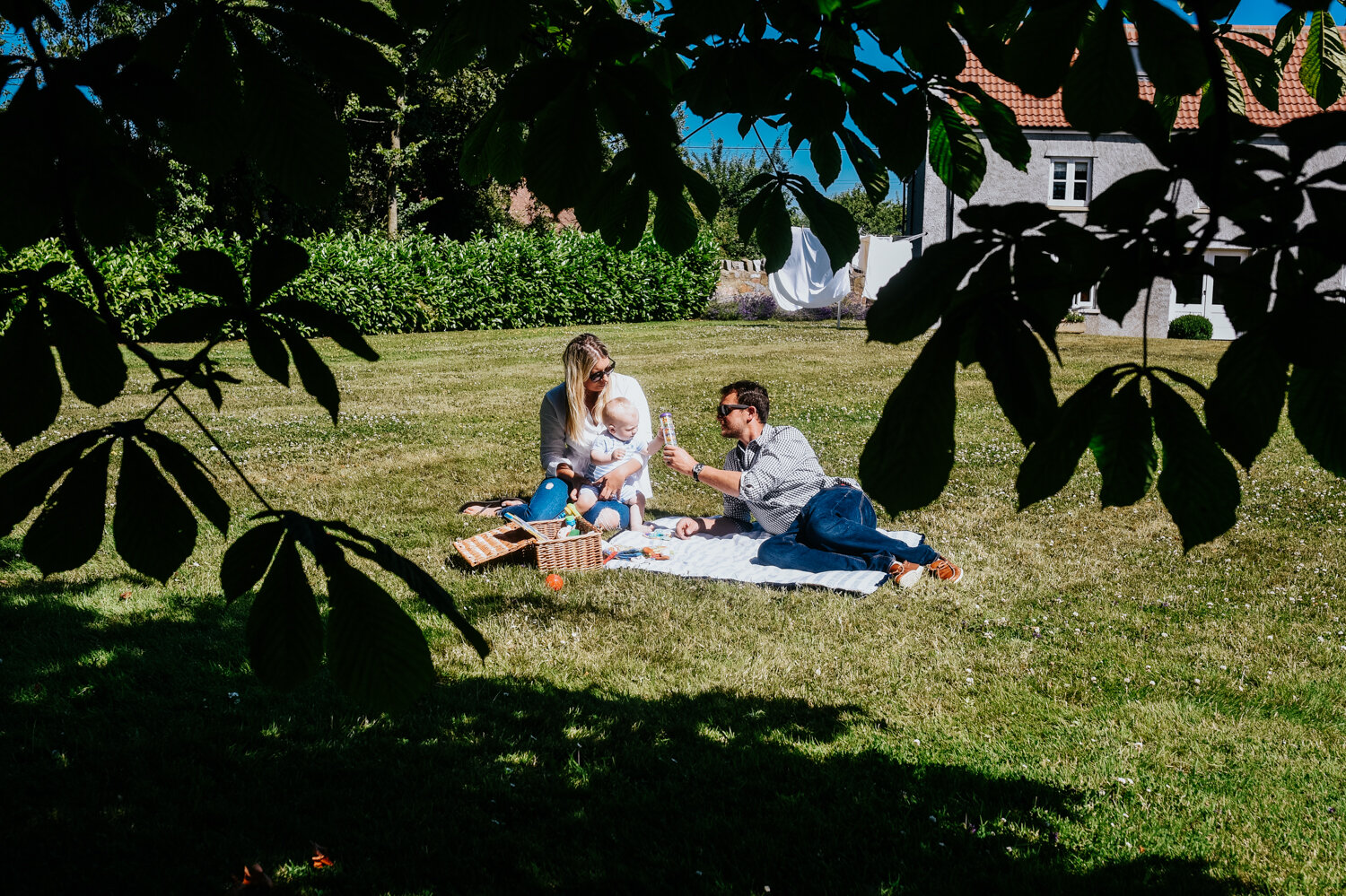  Image is taken through an oak tree as two parents lie on a blanket in their garden in the sun with their baby boy during family photo session in Kingston Seymour, Somerset. 