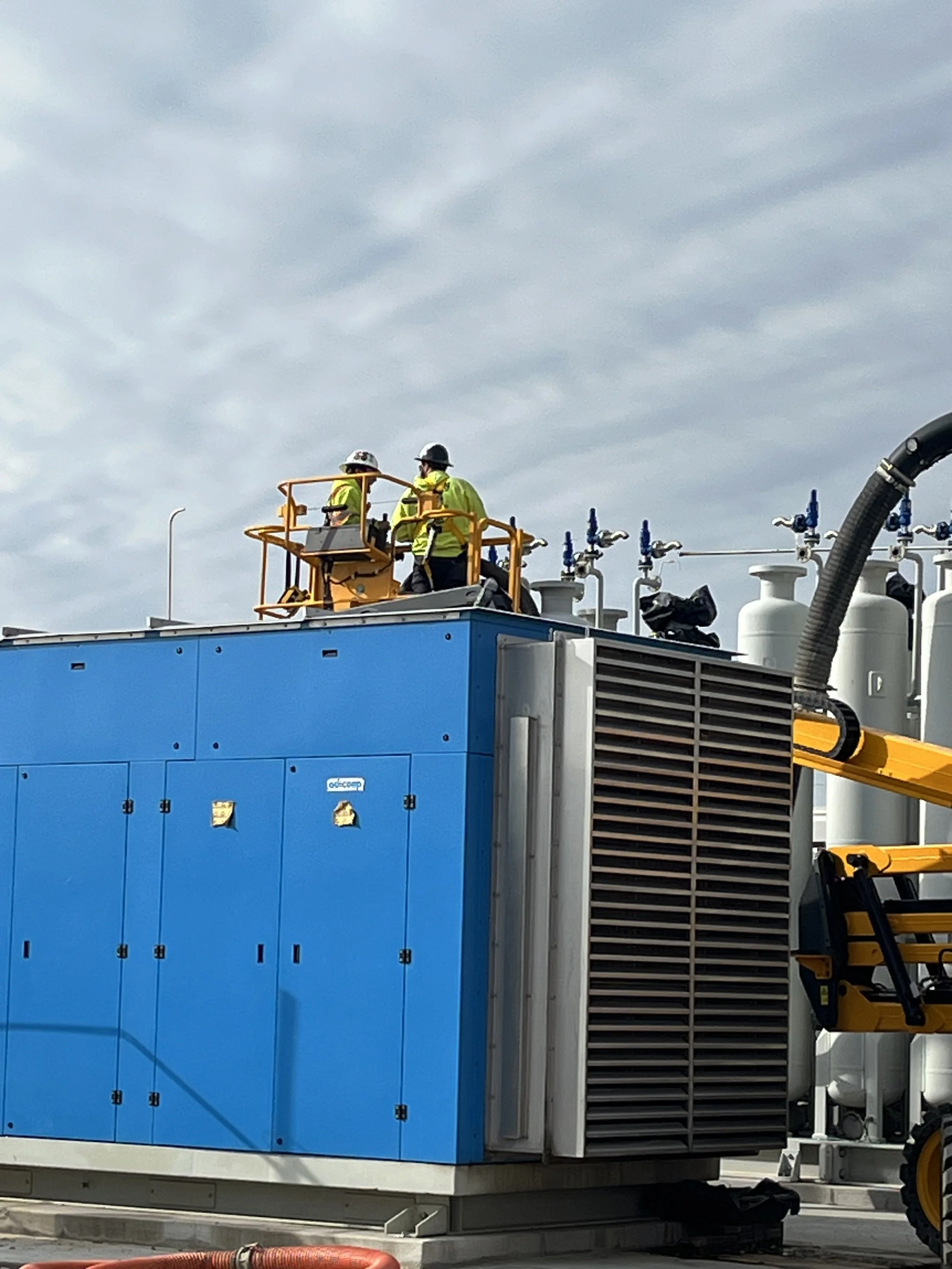 Two workers in safety gear standing in a yellow lift working on electrical equipment on a large blue and white industrial machine, with a cloudy sky in the background.