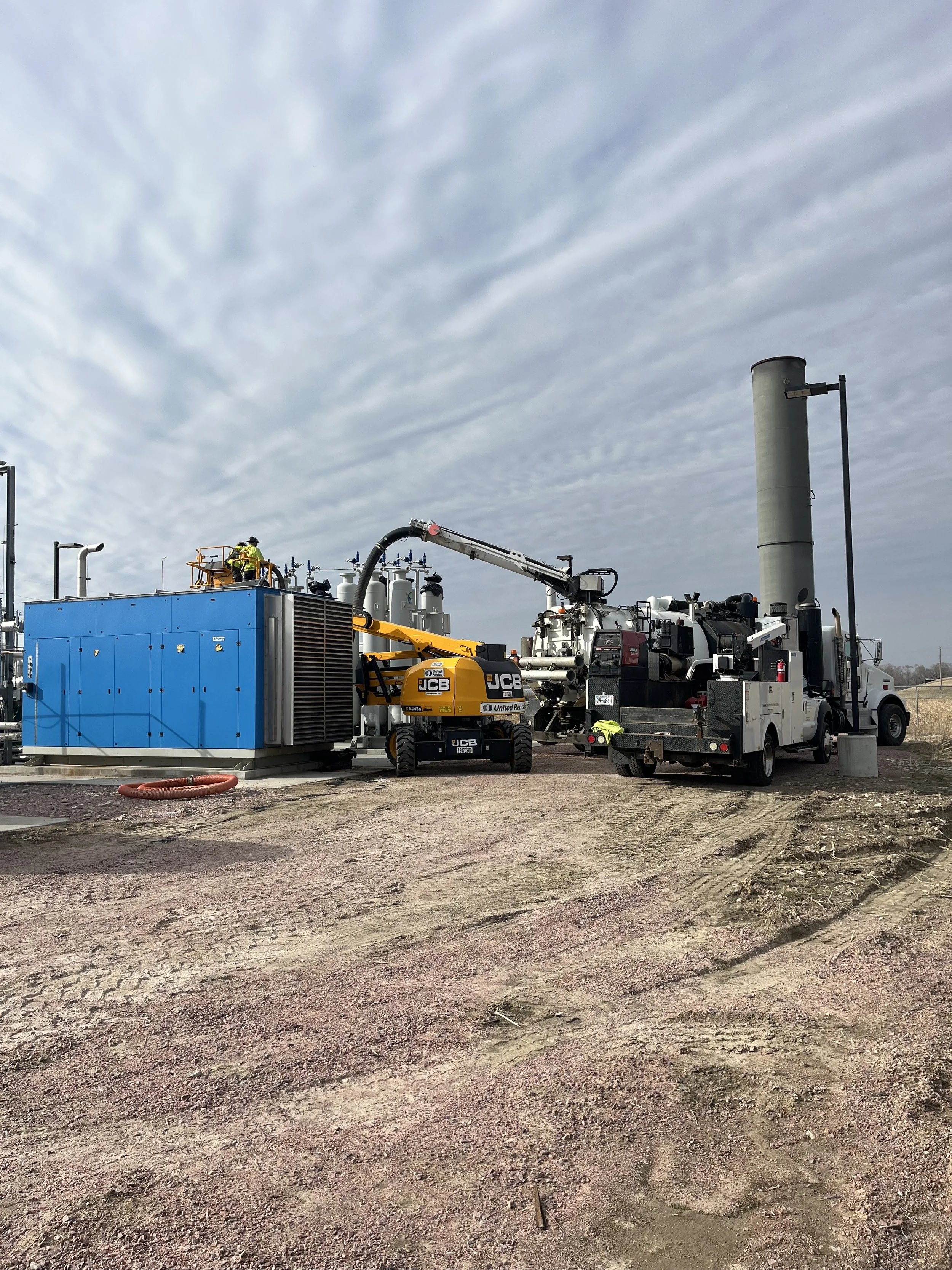 Construction site with heavy machinery, such as a yellow JCB excavator, and workers in safety gear, working on a large industrial piece of equipment against a cloudy sky.