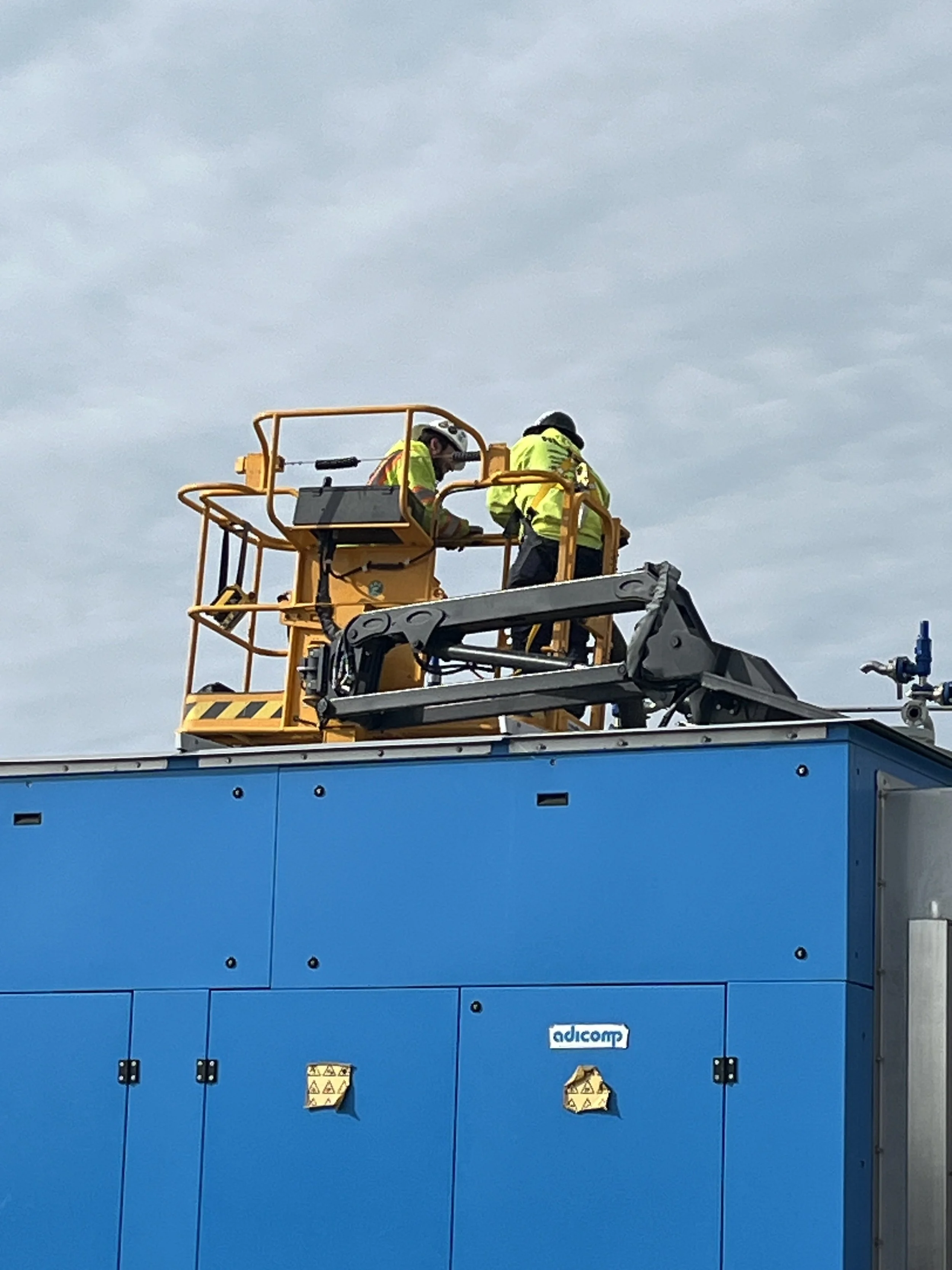 Two workers in yellow safety vests and helmets are operating a construction lift on a building with a blue exterior wall against a cloudy sky.