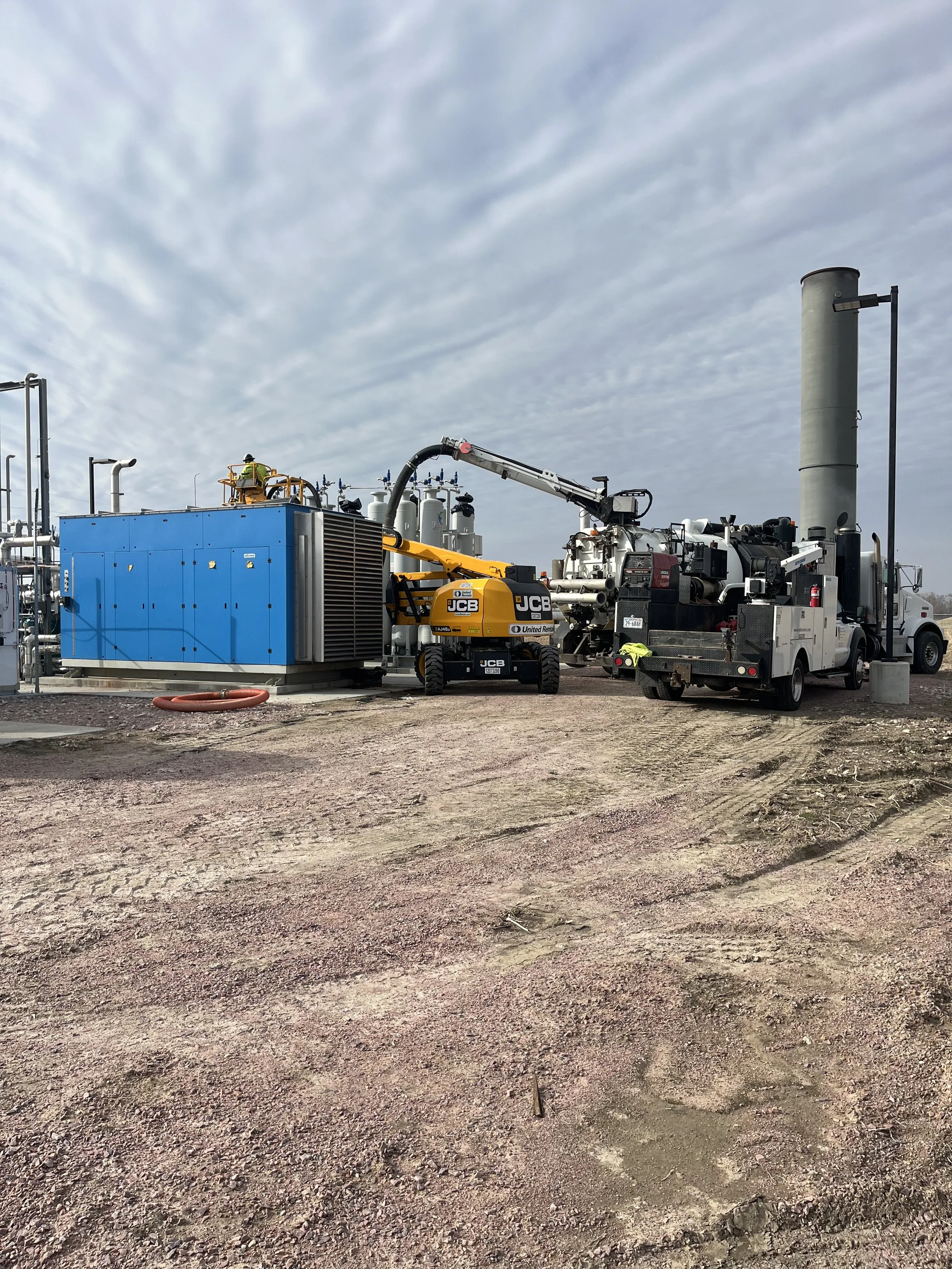 Construction site with heavy machinery, a worker in a yellow safety vest and a hard hat, a blue equipment container, and a large exhaust chimney under a cloudy sky.