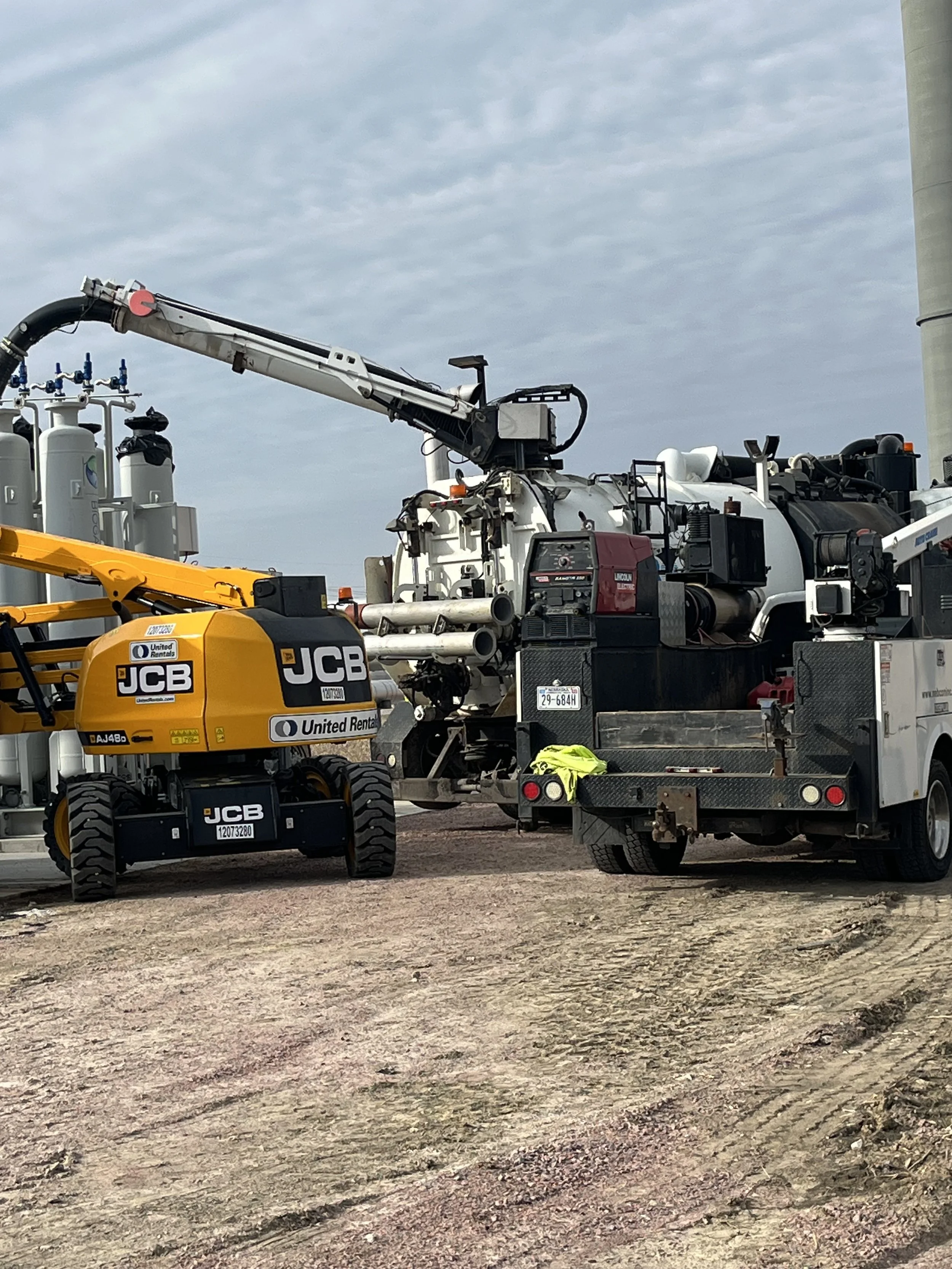 A construction site with various heavy machinery and equipment, including a yellow JCB excavator and a large white machine with a crane arm, set on a dirt ground under a cloudy sky.