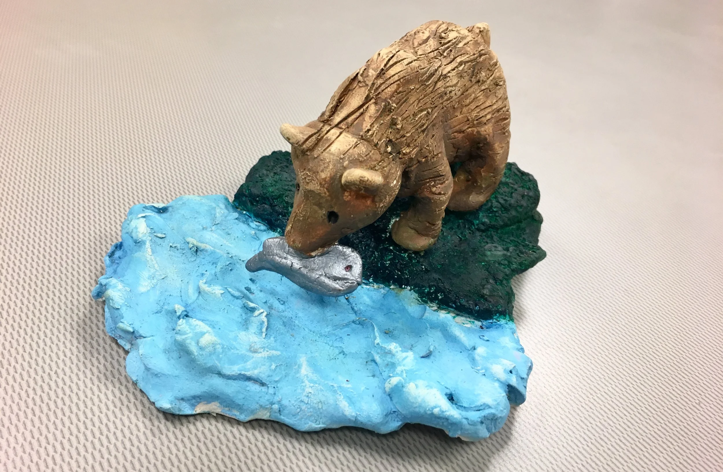 Photo Description: Sculpture of a light brown bear with a fish in its mouth standing on dark green grass with rough blue water.