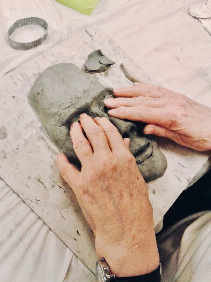 Photo Description: Two hands resting on the eye sockets of a semi-formed sculpture of a face in grey clay.