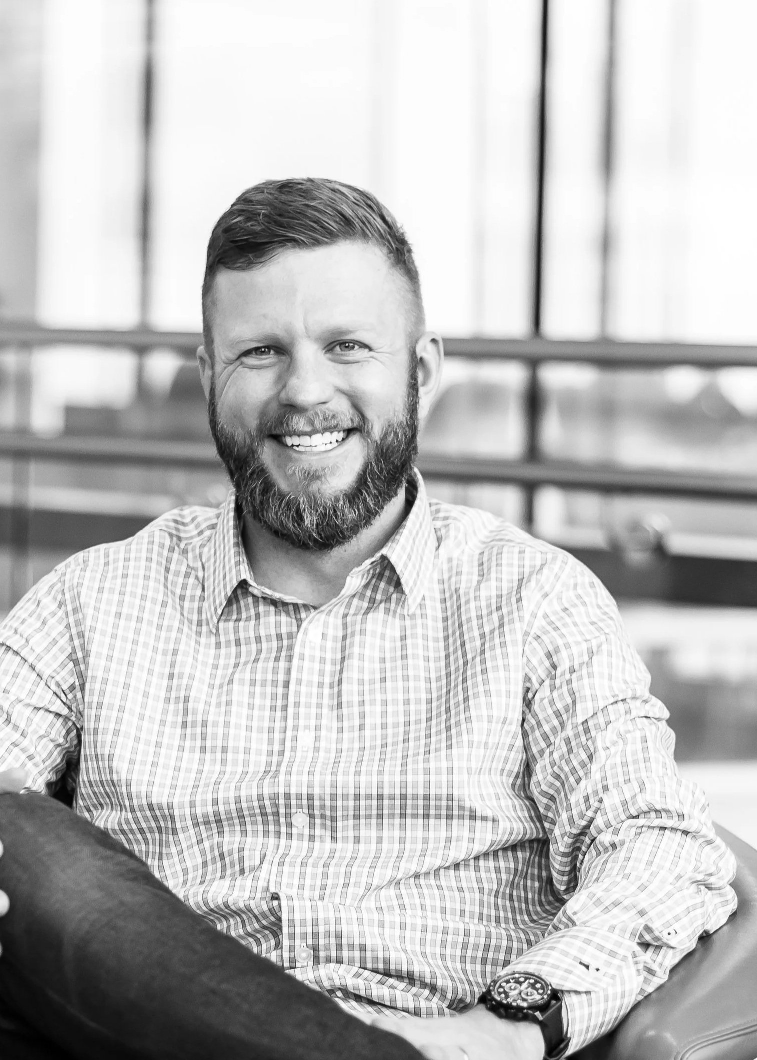 Black and white portrait of a smiling man with a beard, sitting indoors near large windows, wearing a checkered shirt and a wristwatch.