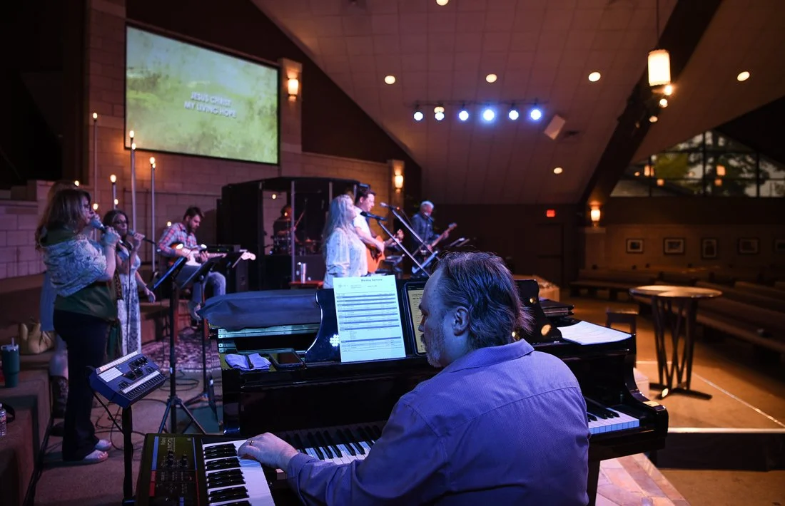 A group of musicians performing on stage with a pianist in the foreground, and a large screen displaying song lyrics in the background.