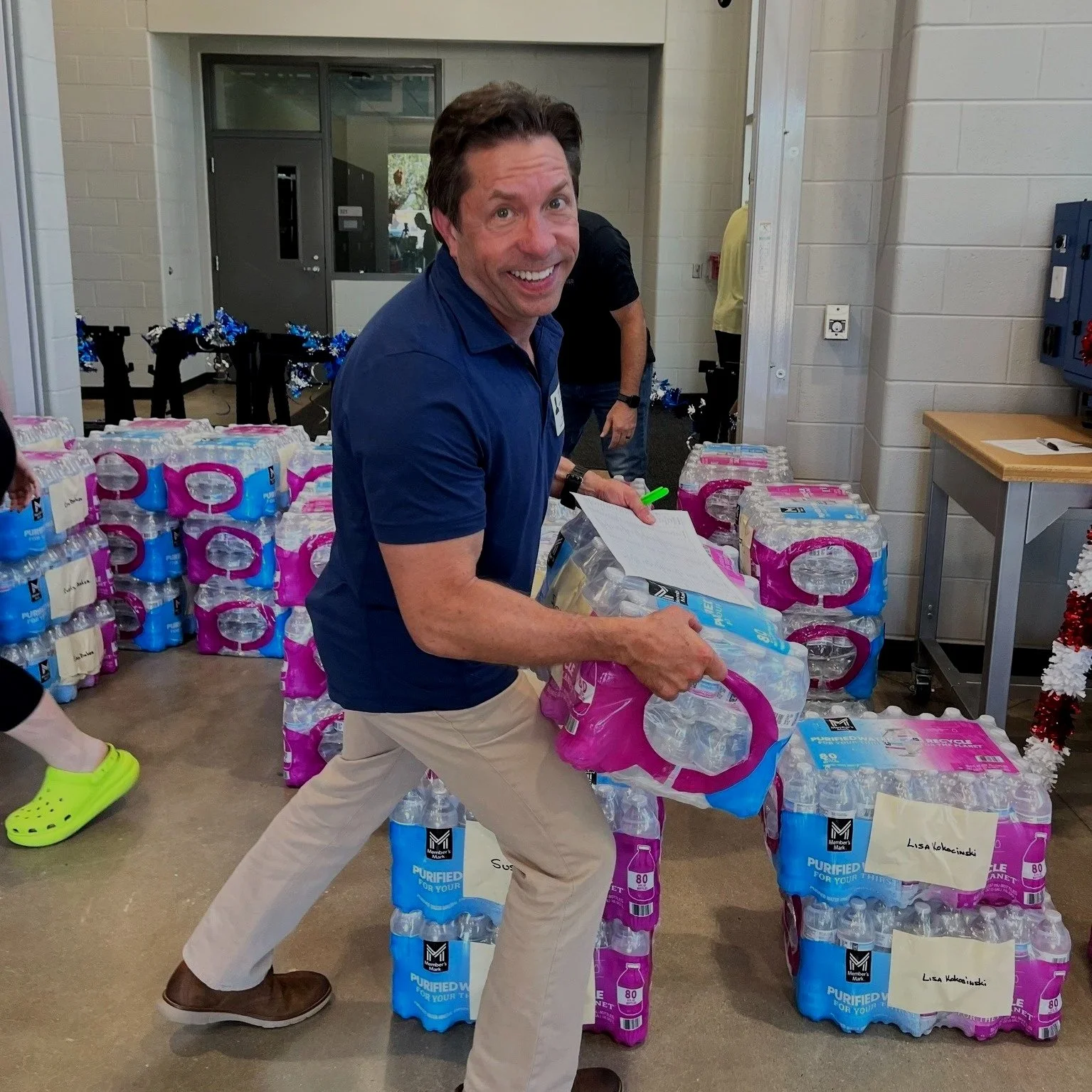 A man smiling and holding a pack of bottled water, surrounded by stacks of bottled water in a room.
