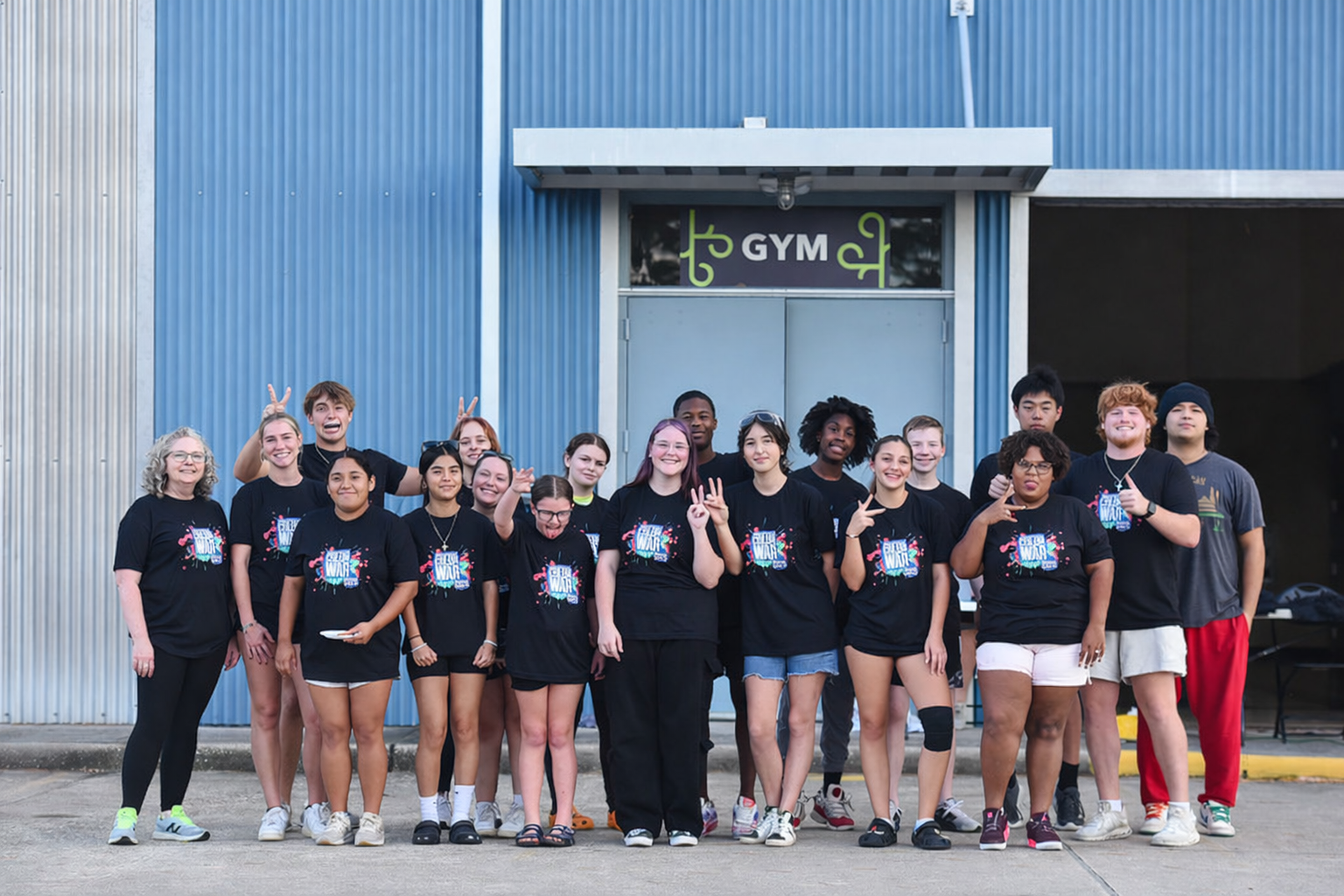 Group of diverse young people standing in front of a blue gym building, posing and smiling for a photo during an outdoor event.