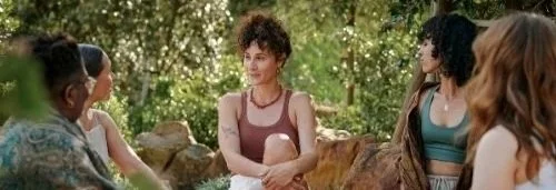 Women sit in a circle in a natural setting, bushes as their backdrop