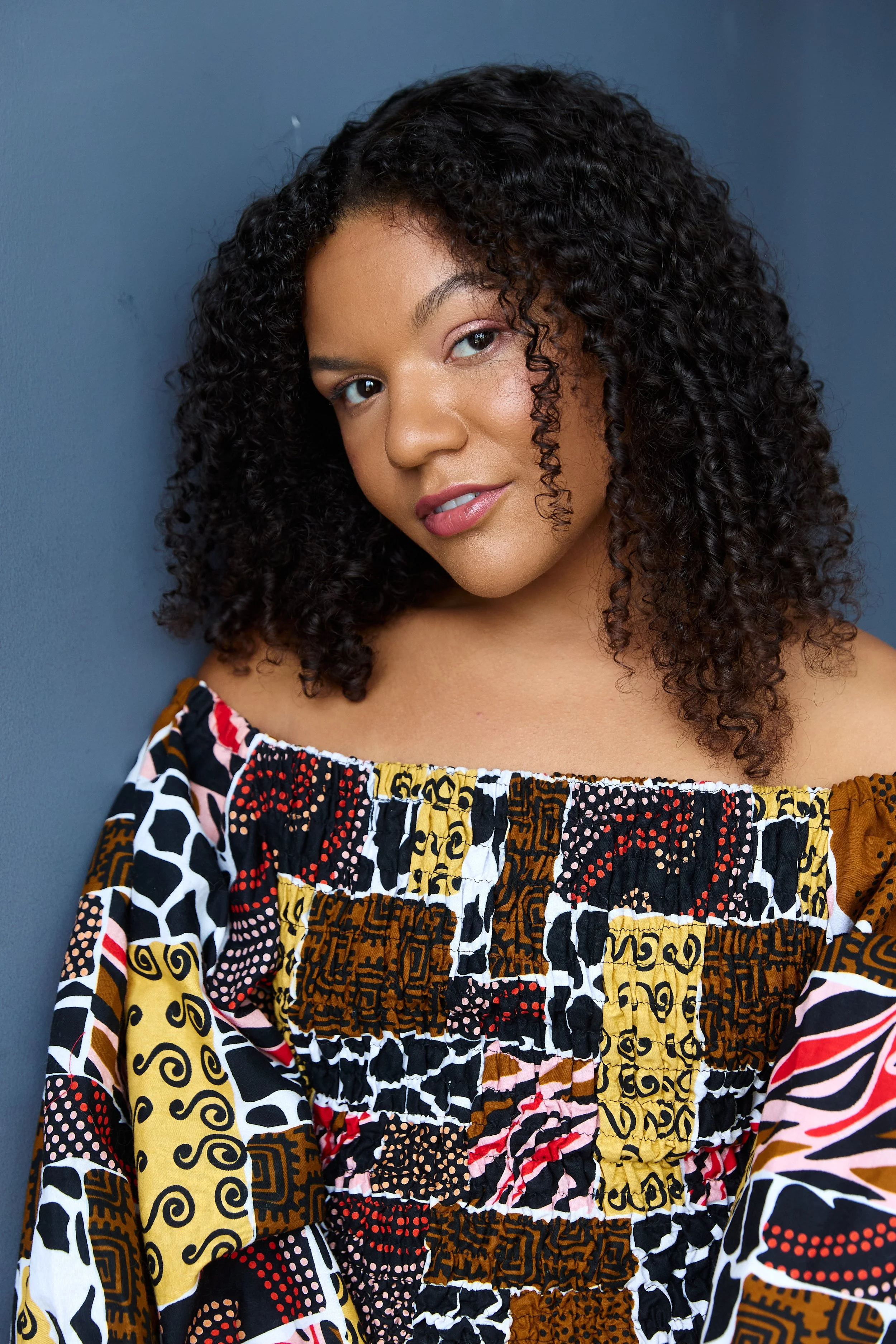 Keira, Biracial, Afro-Caribbean and white Female presenting person is wearing a Vibrant African print shirt, with curly dark hair, pink eyeshadow and a friendly half smile