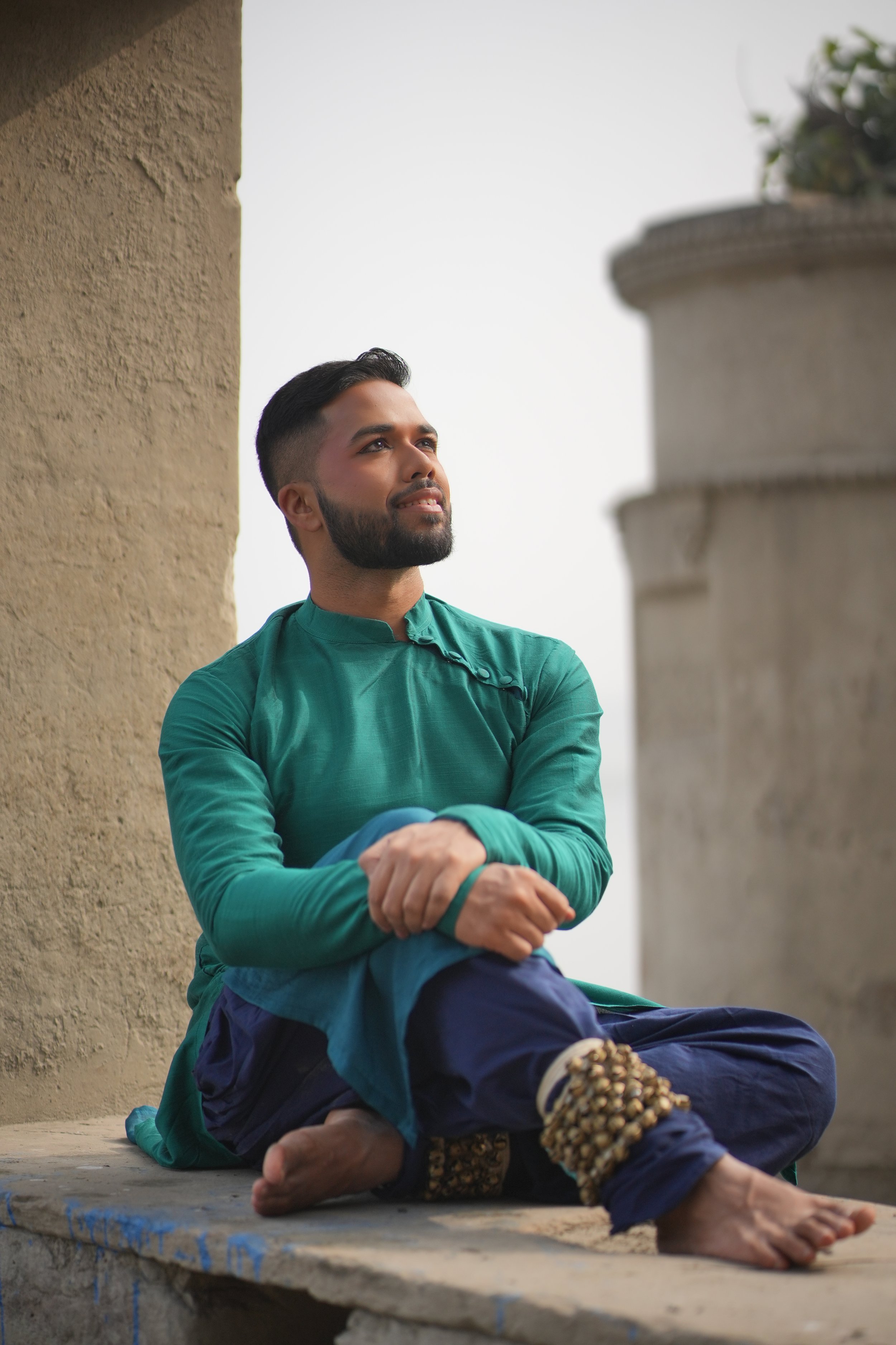 Tanveer, a South Asian man is wearing a blue and green kurta, sitting with his legs crossed, in front of a concrete wall.