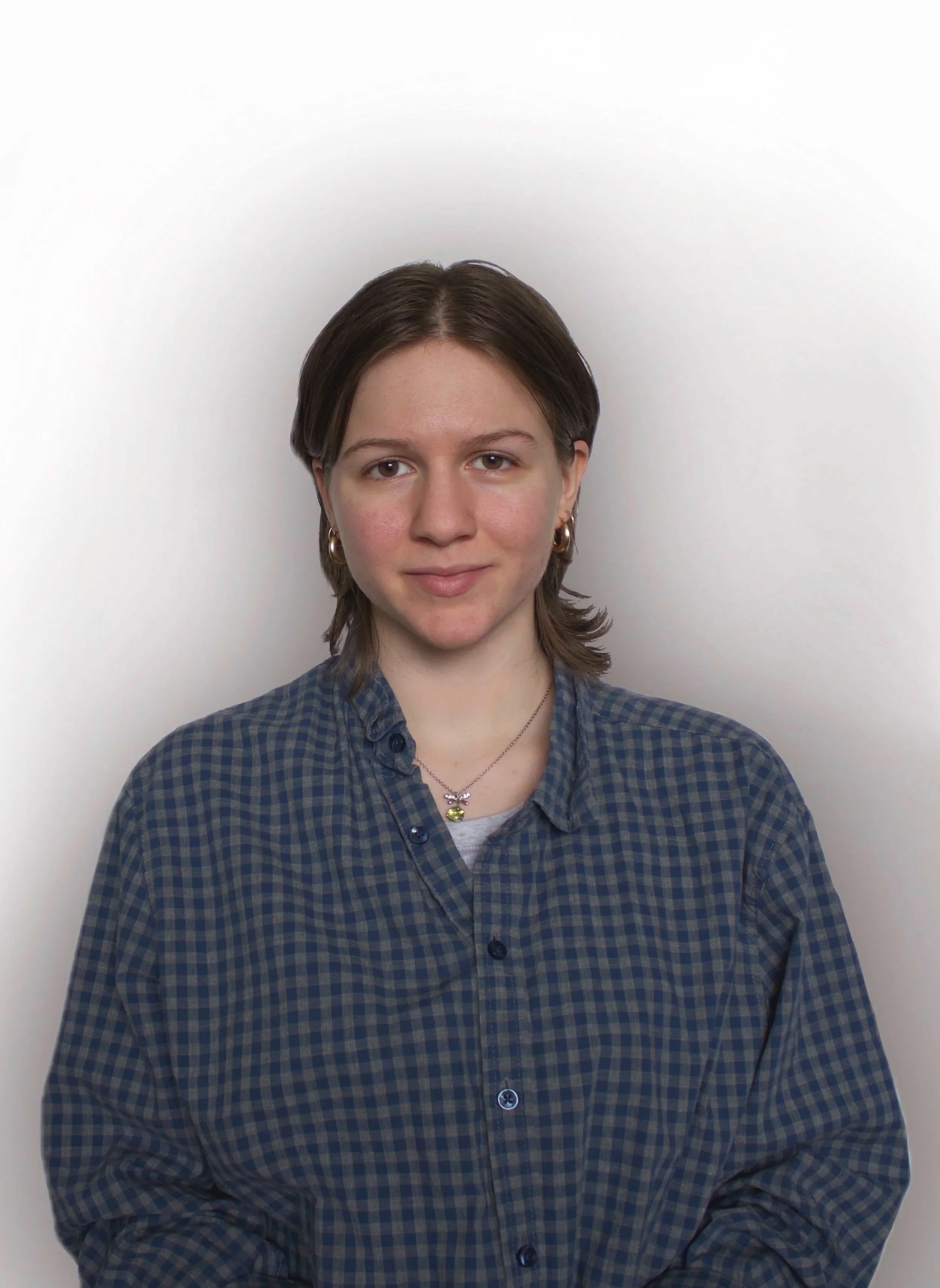 Cass, a white non-binary person with short brown hair wears a blue checkered button-up shirt and smiles to the camera in front of a white background.