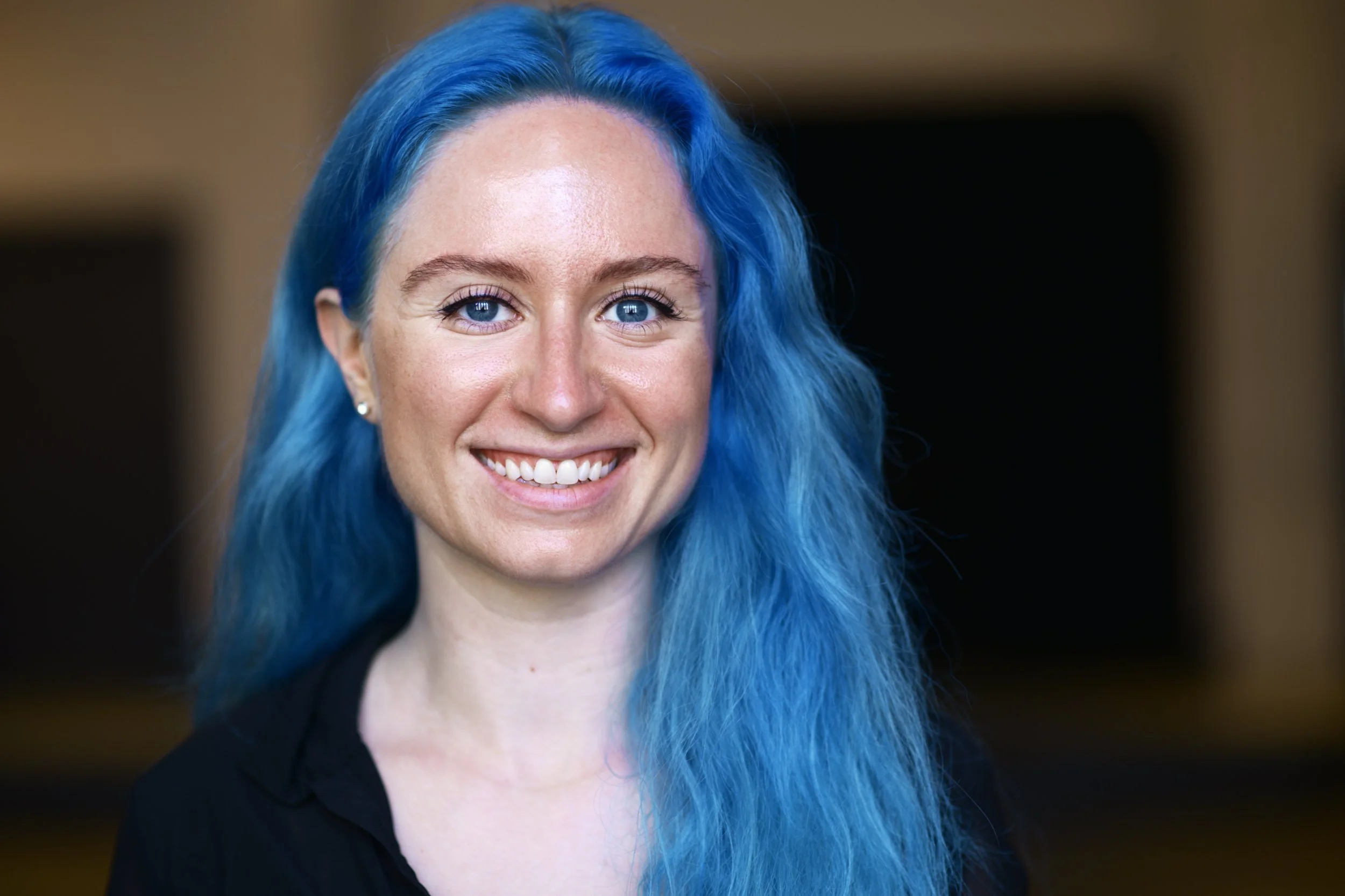 Bri, a Caucasian women, is wearing a black v-neck blouse and smiling inside of a rehearsal studio.