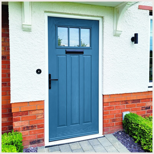 Blue front door with a small window at the top, black handle, and mail slot, set in a white stucco wall with red brick accents, and surrounded by small green bushes.