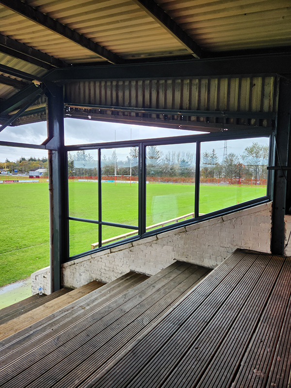 View of a sports field through large windows from a covered seating area with wooden flooring and stairs.