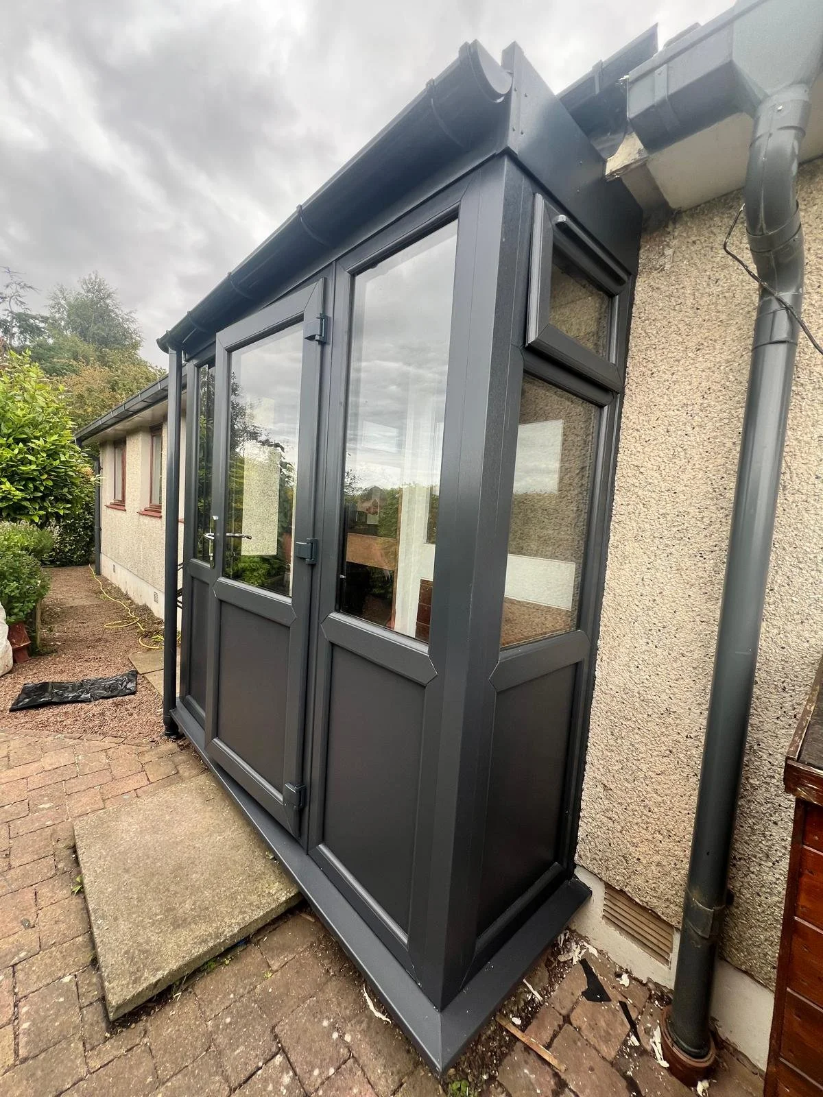 A black-framed glass conservatory attached to the exterior of a house, with a concrete step leading up to the door, surrounded by brick paving and garden plants.